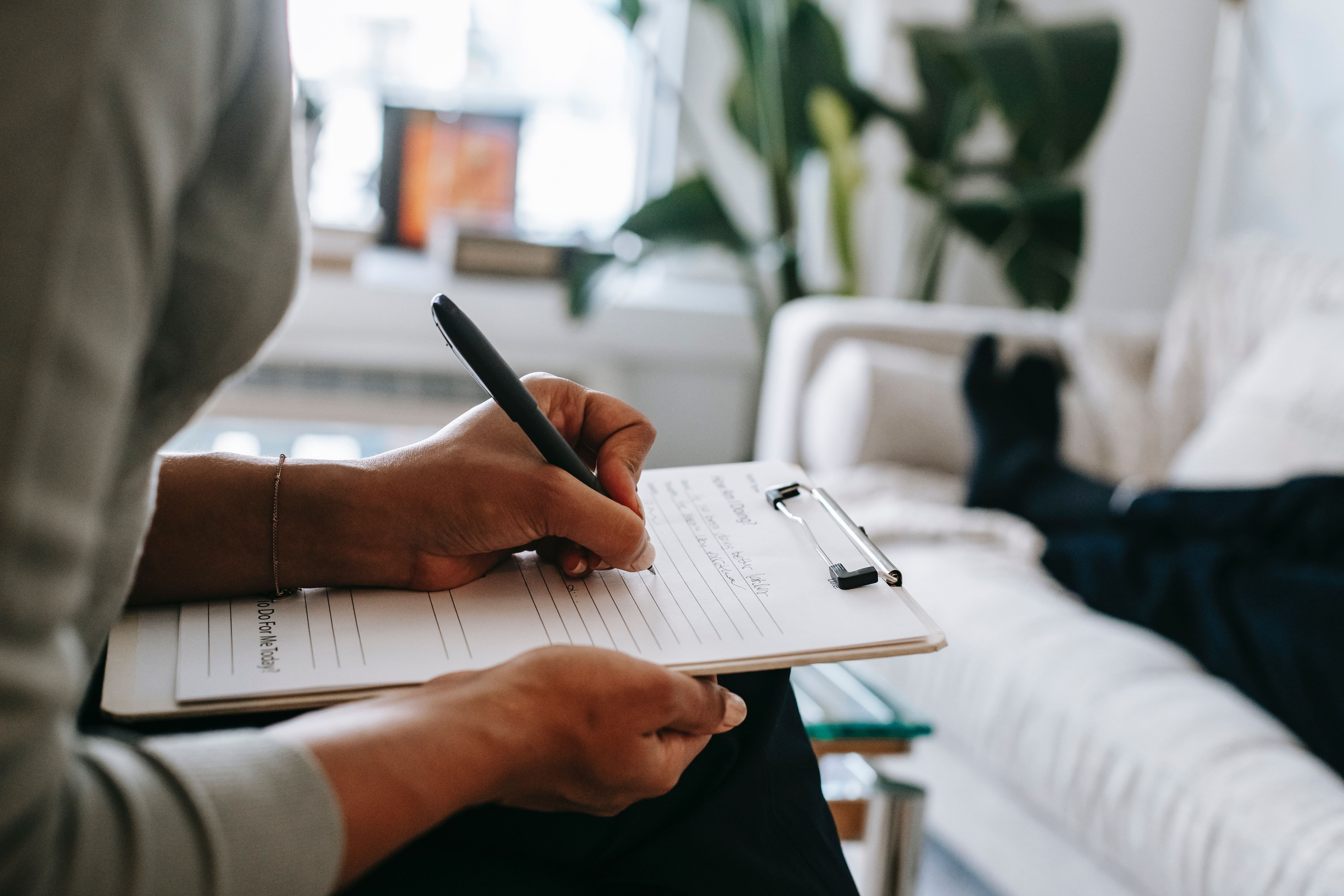 This is an image of a woman sitting on a chair with a notebook and pen in her hand. There is also a couch on which a person is lying down. Seems like a counseling session.