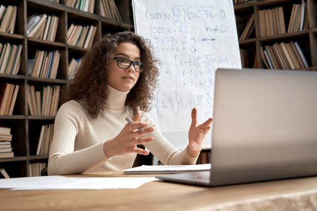 This image shows a woman still in front of a laptop. In the background, there is bookshelves with books and a whiteboard stand with some algebra trigonometry question and working. The woman appeared to be having a conversation with someone on the laptop screen. She has a pen in her right hand and a notepad in front of her.