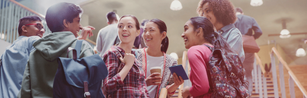 This is a rectangular image capturing a group of college students engaged in conversation on a stairway. There are four girls and two boys, all looking at each other with interest and attentiveness. The atmosphere is lively and sociable, with the students engrossed in discussion. The stairway provides a casual and comfortable setting for their interaction, fostering a sense of camaraderie and academic community among the group.