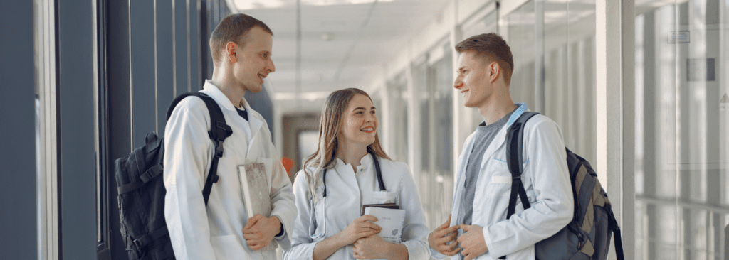 Group of people standing in a hallway smiling. They are wearing white doctor jackets. The hallway is full of windows.