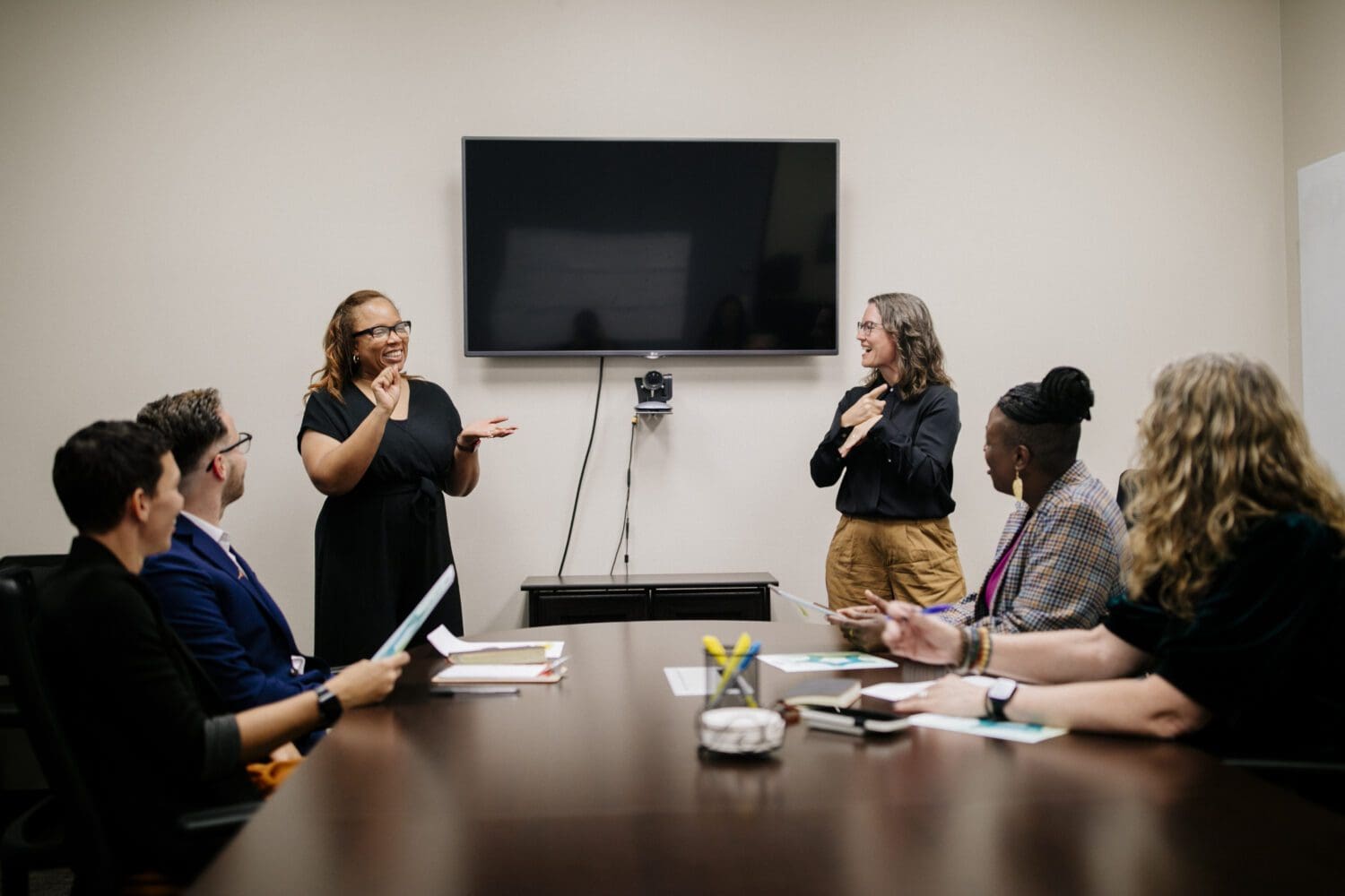 A group of seven people are in a professional meeting room. Two women stand at the front of the room, smiling and communicating in American Sign Language (ASL). One woman, wearing a black dress and glasses, is signing while the other, in a black blouse and tan pants, signs back. Behind them is a large flat-screen TV mounted on the wall above a webcam. Five others sit around a long conference table, watching and engaging with the presenters. The table has notebooks, pens, and documents, and the mood appears collaborative and positive.
