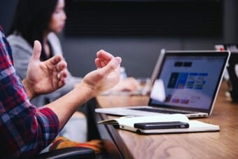 The image depicts a professional setting with two people at a conference table. In the foreground, a person is gesturing with their hands while speaking. This person wears a plaid shirt. In front of them is a closed notebook with a smartphone placed on top. A laptop is open on the table, displaying a colorful webpage or application interface.