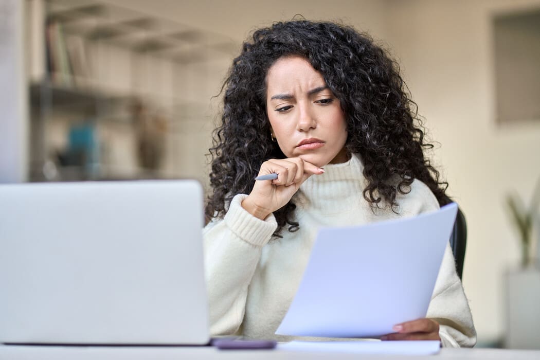 A person with long, curly dark hair is seated at a desk, focusing intently on a document they are holding in their left hand. They appear thoughtful, resting their chin on their right hand and holding a pen. They are wearing a light-colored, cozy turtleneck sweater. In front of them is an open laptop.