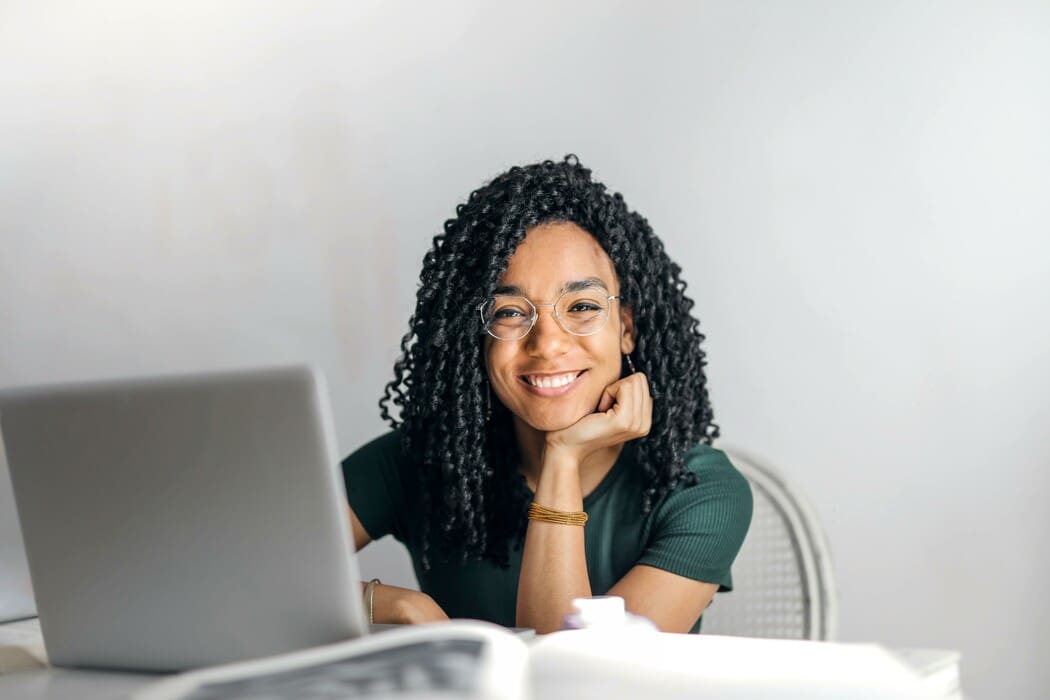 The image shows a person with curly hair and glasses sitting at a table with a laptop in front of them. They are wearing a dark green shirt and are smiling, resting their chin on one hand. The background is an off-white color, giving the scene a bright and airy feel. 