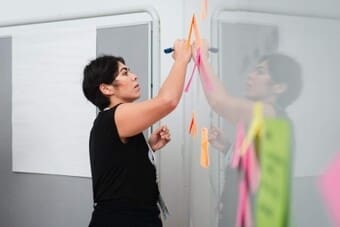 The image shows a person with short dark hair writing on a transparent glass or acrylic board using a dark-colored marker. The individual is positioned on the left side of the image, wearing a black sleeveless top. On the board, several colorful sticky notes in shades of pink, orange, and green are attached, some of which are blurred in the foreground and mirror the action