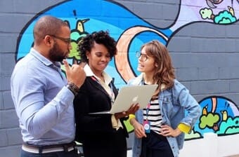 The image shows three people standing in front of a colorful mural painted on a brick wall. On the left, a person wearing a light blue shirt gestures with their hand while engaging in conversation. In the center, a person with curly hair, wearing a black blazer, holds an open laptop, appearing to show something on the screen to the others. On the right, a person wearing glasses, a striped shirt, and a denim jacket, looks at the laptop attentively with one hand on their hip. 