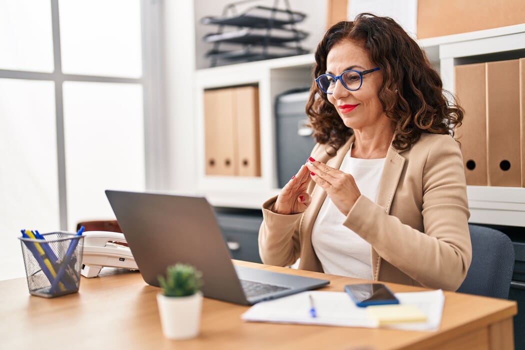 The image shows a woman seated at a desk in an office setting, using a laptop. She has shoulder-length curly hair and is wearing a beige blazer over a white blouse. She is smiling and appears engaged in a sign language conversation. The desk is light wood and holds several items including a laptop, a smartphone, and a pen holder with pens.