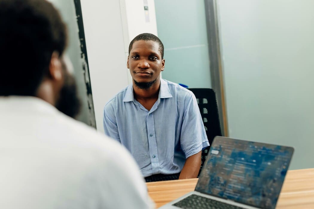 The image shows two people sitting across from each other at a wooden table. In the foreground, there is a blurred view of the back of one person's head and shoulder, and a laptop screen displaying blurred content. The focus is on a man with a short haircut and facial hair, wearing a light blue shirt, who is sitting across the table and looking towards the camera.