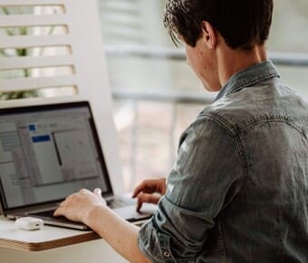 The image depicts a person sitting at a desk working on a laptop. The individual is wearing a denim shirt and has short hair. The desk is white and has a minimalistic design, with a small electronic device resting on it beside the laptop. 