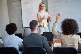 The image shows a classroom or seminar setting where a person stands at the front, facing three seated individuals. The person at the front, with light hair, is wearing glasses and a sleeveless top. They appear to be speaking or presenting, with their hands gesturing. Behind them, a whiteboard is visible with various colored markers and diagrams drawn on it. Among the three attendees, two have raised their hands.