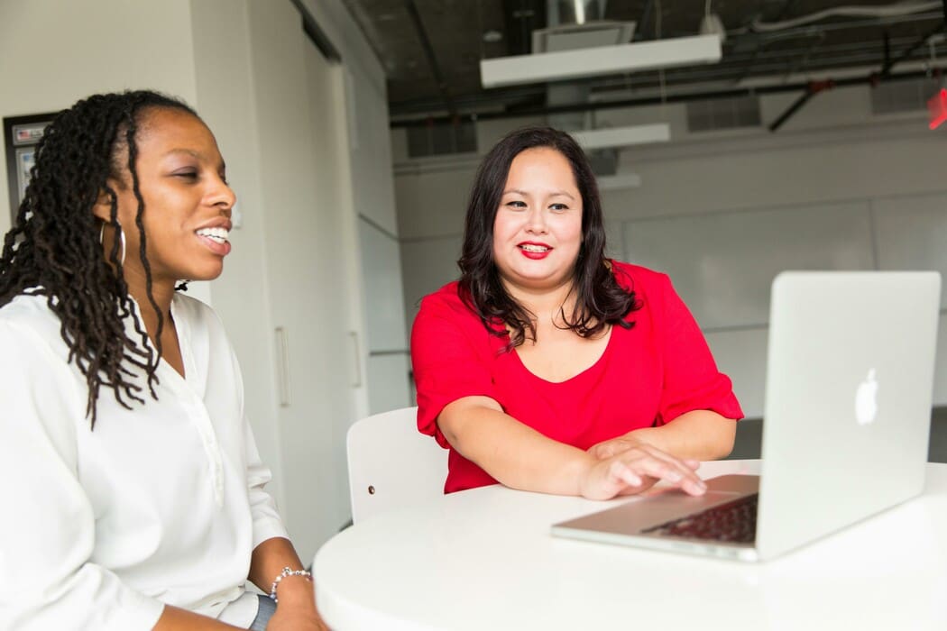 Two colleagues sit at a round table, discussing work while viewing a laptop in a bright office.