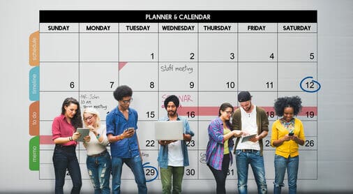 The image depicts seven people standing in front of a large planner and calendar. The calendar features a weekly grid layout with days of the week labeled at the top, from Sunday to Saturday. Boxes for each day show numbers representing dates.The individuals in front, diverse in appearance, are engaging with various electronic devices like tablets and laptops, appearing focused and collaborative. 