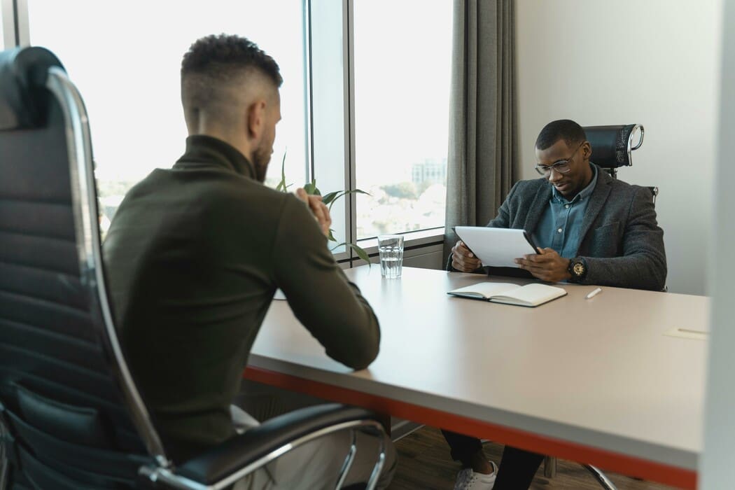 The image depicts a professional setting with two individuals seated across from each other at a desk. The person on the left is viewed from behind and is wearing a dark suit, sitting on a black chair. The individual on the right is wearing glasses, a blazer, and a blue shirt, engrossed in reading a document or tablet. A glass of water, an open notebook, and a pen are neatly arranged on the desk between them