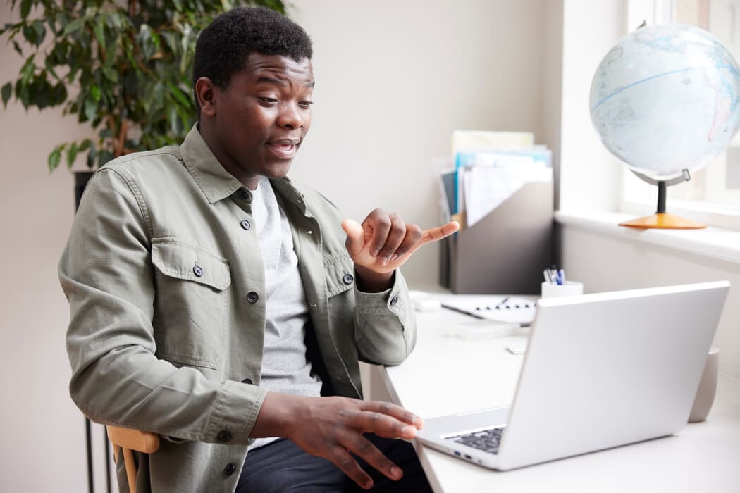 The image shows a person sitting at a desk, engaged in a video call on a laptop. They are wearing a light gray jacket over a white shirt. The person is using sign language, possibly emphasizing a point. The background includes a potted plant and a globe on a stand by the window, with natural light filtering in. 
