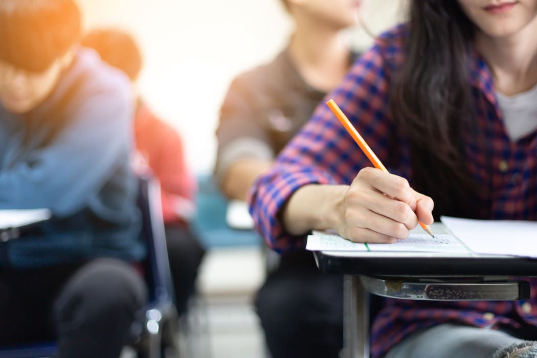 The image shows a classroom setting where students are seated at desks taking an exam. In the foreground, one student with long dark hair, wearing a red and blue checkered shirt, is focused on writing with a pencil on an exam sheet placed on a desk. The desk has a black metal frame with a wooden writing surface
