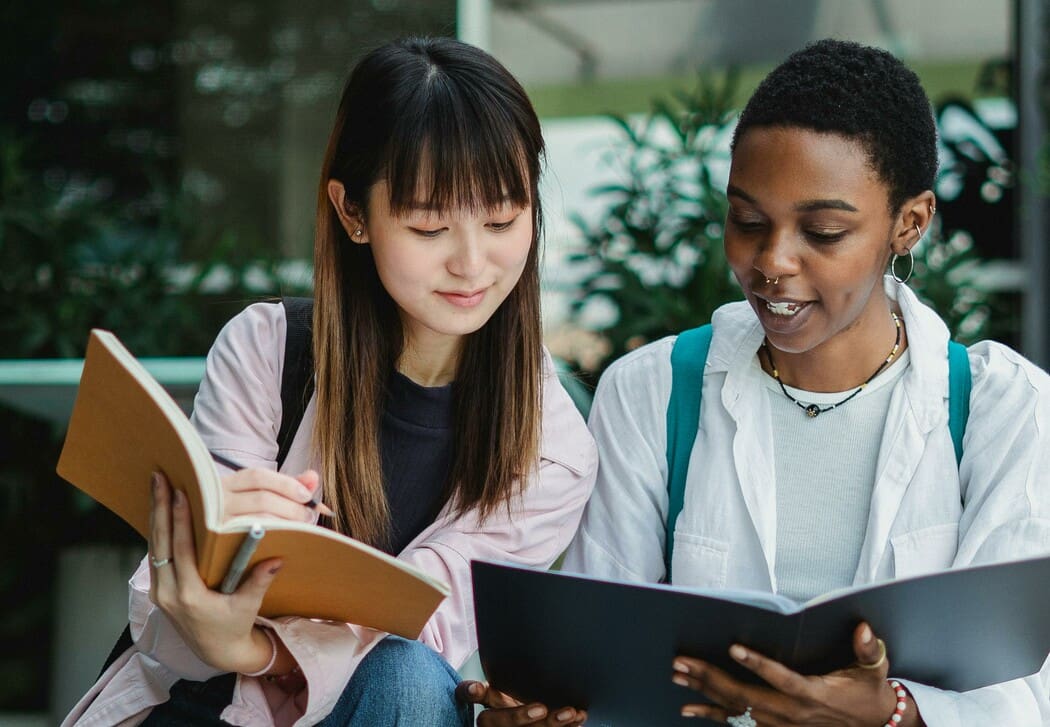 The image shows two people sitting closely together outdoors, engaged in studying. The person on the left has long dark hair and is holding an open notebook with blank lined pages. They are dressed in a light pink jacket over a black shirt. The person on the right has short hair and is holding an open folder or book with dark pages. They are wearing a white shirt over a light top and a necklace.