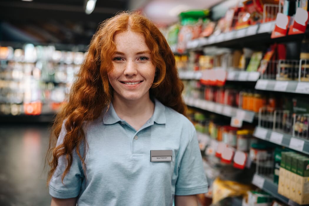 Smiling grocery store employee with long red hair standing in an aisle.