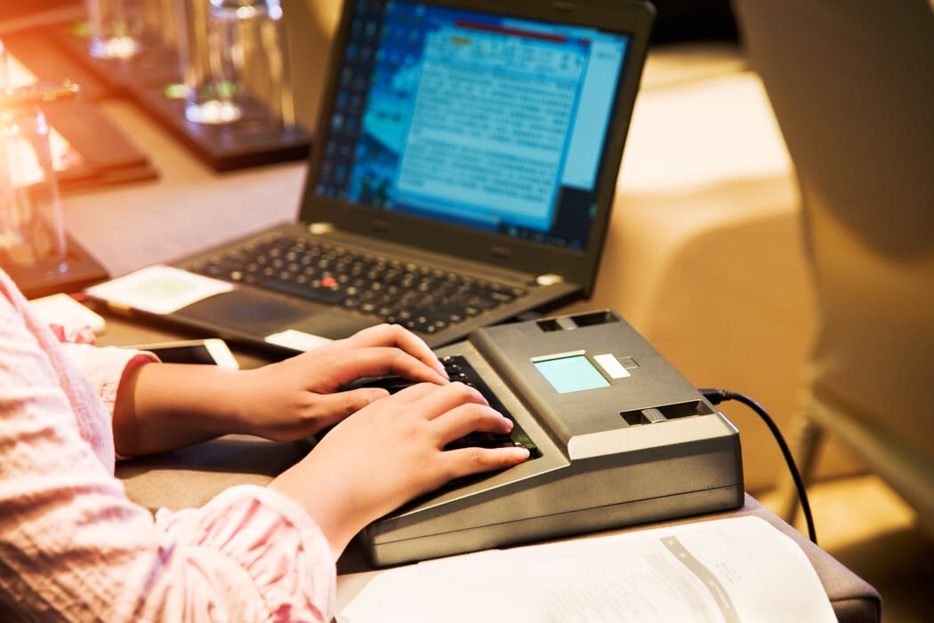 The image shows a person using a stenograph machine, positioned on a desk next to a laptop. The laptop screen displays a blurred interface with unreadable text. The stenograph machine is gray and has a display screen. The person is typing on the stenograph with both hands, suggesting transcription or captioning activity. 