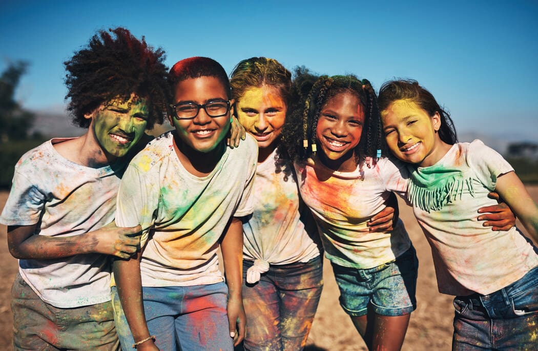 The image shows a group of five children standing closely together outdoors, smiling brightly. They are covered in colorful powder, suggesting they are participating in a celebration or festival similar to Holi. The powder has left vibrant hues of green, red, yellow, and orange on their faces, hair, and clothes. The children are wearing casual clothing, mostly white T-shirts and shorts, which accentuate the colors smeared on them. 
