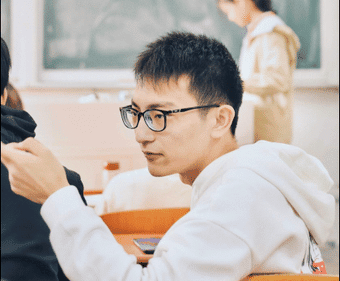 The image shows a classroom scene with a focus on a student wearing glasses and a white hoodie. The student is seated and appears engaged, holding a pencil or pen in one hand. 