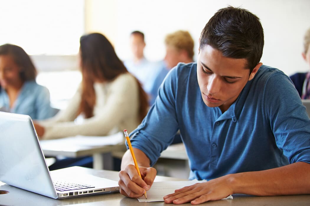 The image depicts a classroom setting with several students seated at desks. In the foreground, a male student with short dark hair is focused on writing on a piece of paper. He is wearing a long-sleeved blue shirt and holds a pencil in his right hand. A silver laptop is open on the desk beside him. 