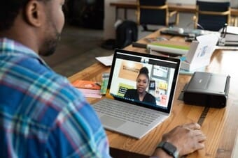The image depicts a man sitting at a wooden table, engaged in a video call on a laptop. He is wearing a plaid shirt and a watch on his left wrist. The laptop is set on the table, displaying a woman speaking in the video chat window. The table is cluttered with various items, including a printer, a stack of papers, and some colorful notebooks. 