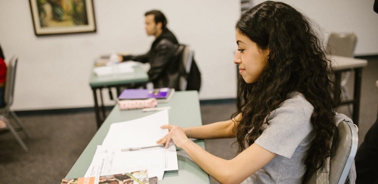 The image shows a classroom setting with two students sitting at desks. In the foreground, a student with long, curly dark hair is seated at a green desk, focusing on papers in front of her. She is wearing a light gray shirt and has a pen resting on the paper.