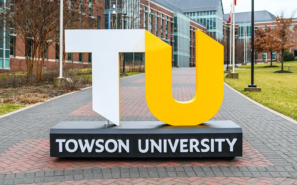 A large freestanding sculpture displays the letters “TU” — the “T” in white and the “U” in bright yellow — representing Towson University. The sculpture sits on a black base with “TOWSON UNIVERSITY” written in white text. It stands on a paved walkway lined with brick buildings, trees, and lamp posts on a university campus.