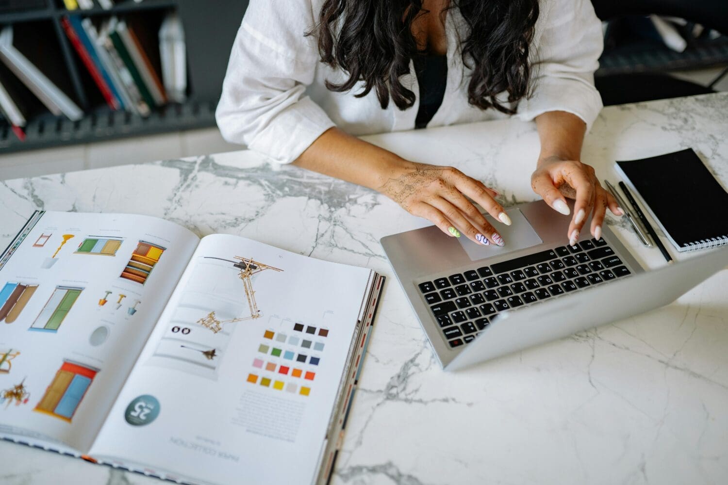 A person sits at a marble desk working on a laptop beside an open design book showing color swatches and sketches. Their hands, decorated with henna and colorful nails, rest on the keyboard. A notebook and pen lie nearby, suggesting a creative or design-focused workspace.