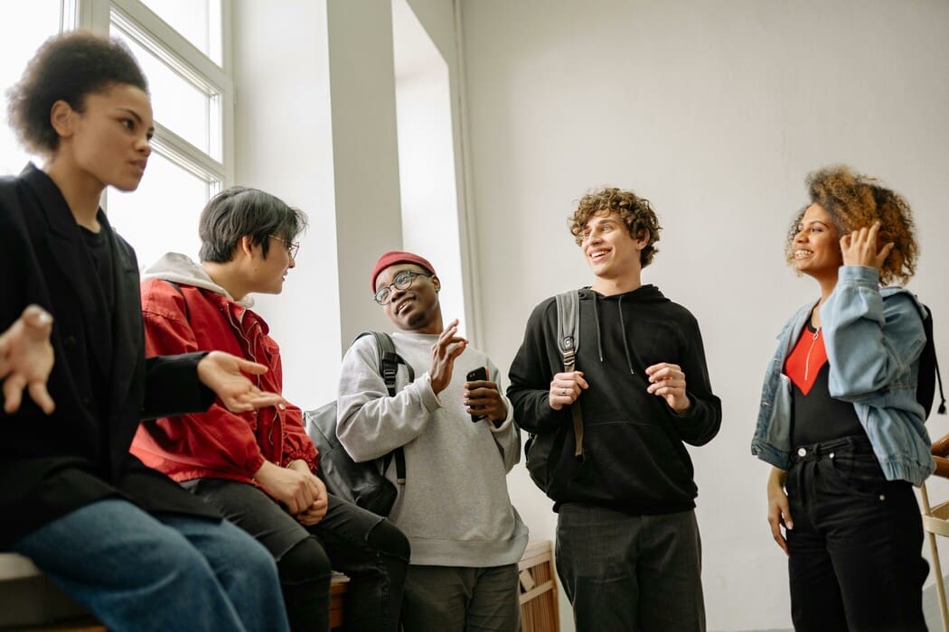 The image depicts five people casually engaging in conversation in a well-lit indoor setting. On the left, a person with curly hair gestures with their hands while seated on a ledge. Next, a person in a red jacket and glasses smiles, facing the group. The third individual, wearing a gray hoodie and red beanie, is holding a smartphone and making a conversational gesture. Beside them, a person in a black hoodie laughs, wearing a backpack. On the far right, another person with curly hair touches their ear, wearing a denim jacket over a red shirt.