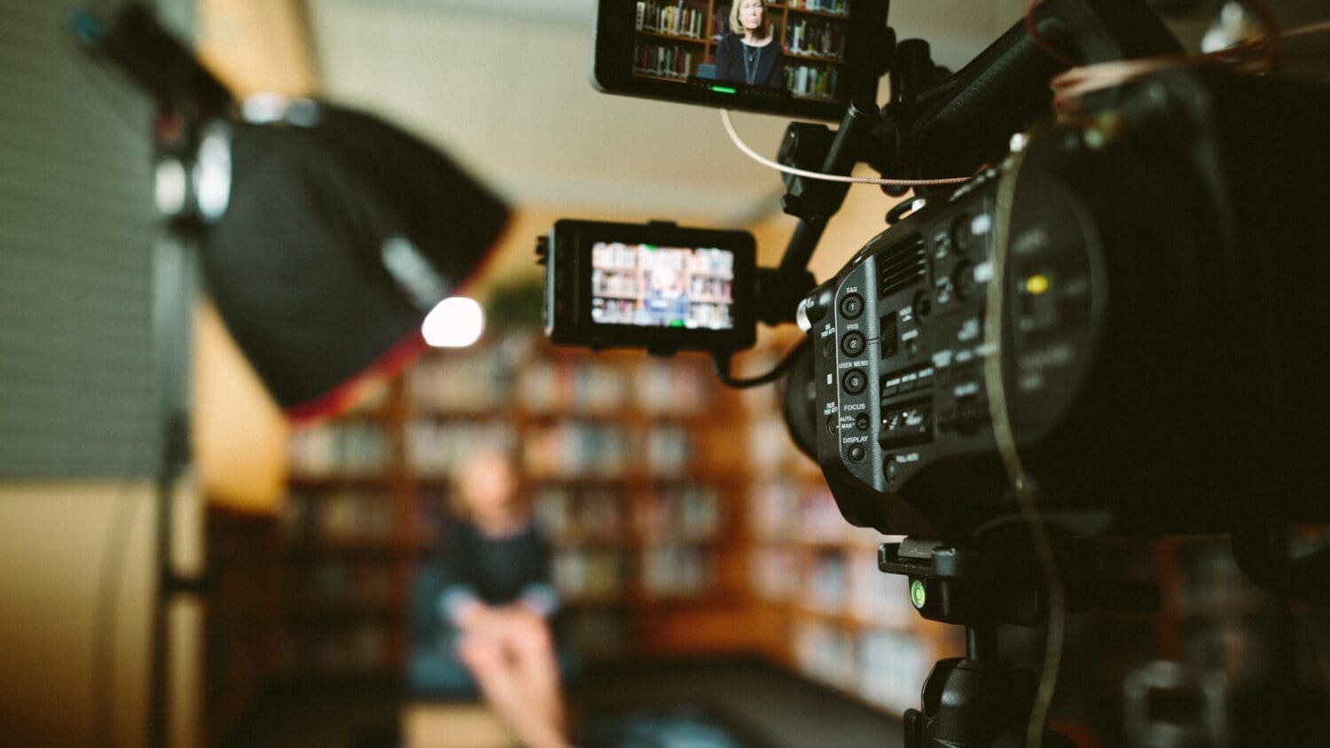 The image depicts a recording setup focused on a videocamera in a library setting. In the foreground, the camera is prominently displayed on the right, mounted on a tripod. Its lens is pointed toward a seated person in the background, who is blurred, indicating they are the subject of the recording. The background shows bookshelves filled with colorful books, slightly out of focus.