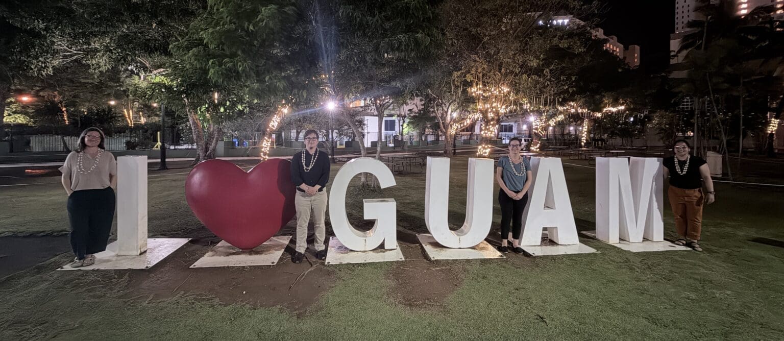 Cuatro personas posan por la noche junto a un gran cartel que dice 'Yo amo GUAM' en un parque con árboles y luces de cadena.